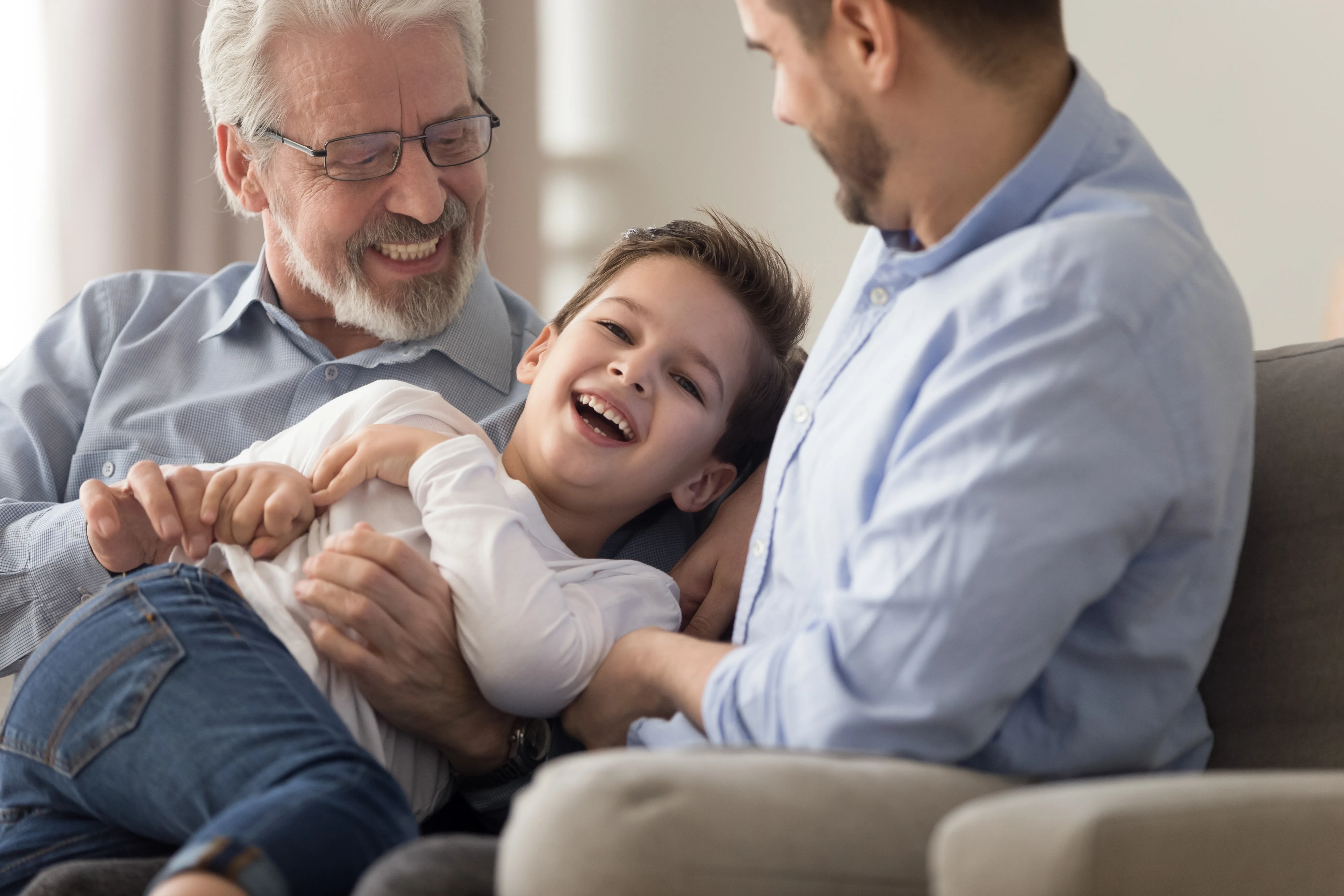 Close up focus on boy have fun with grandpa and father play on couch in living room, multi generational relatives men spend free time together at home tickling laughing family leisure activity concept
