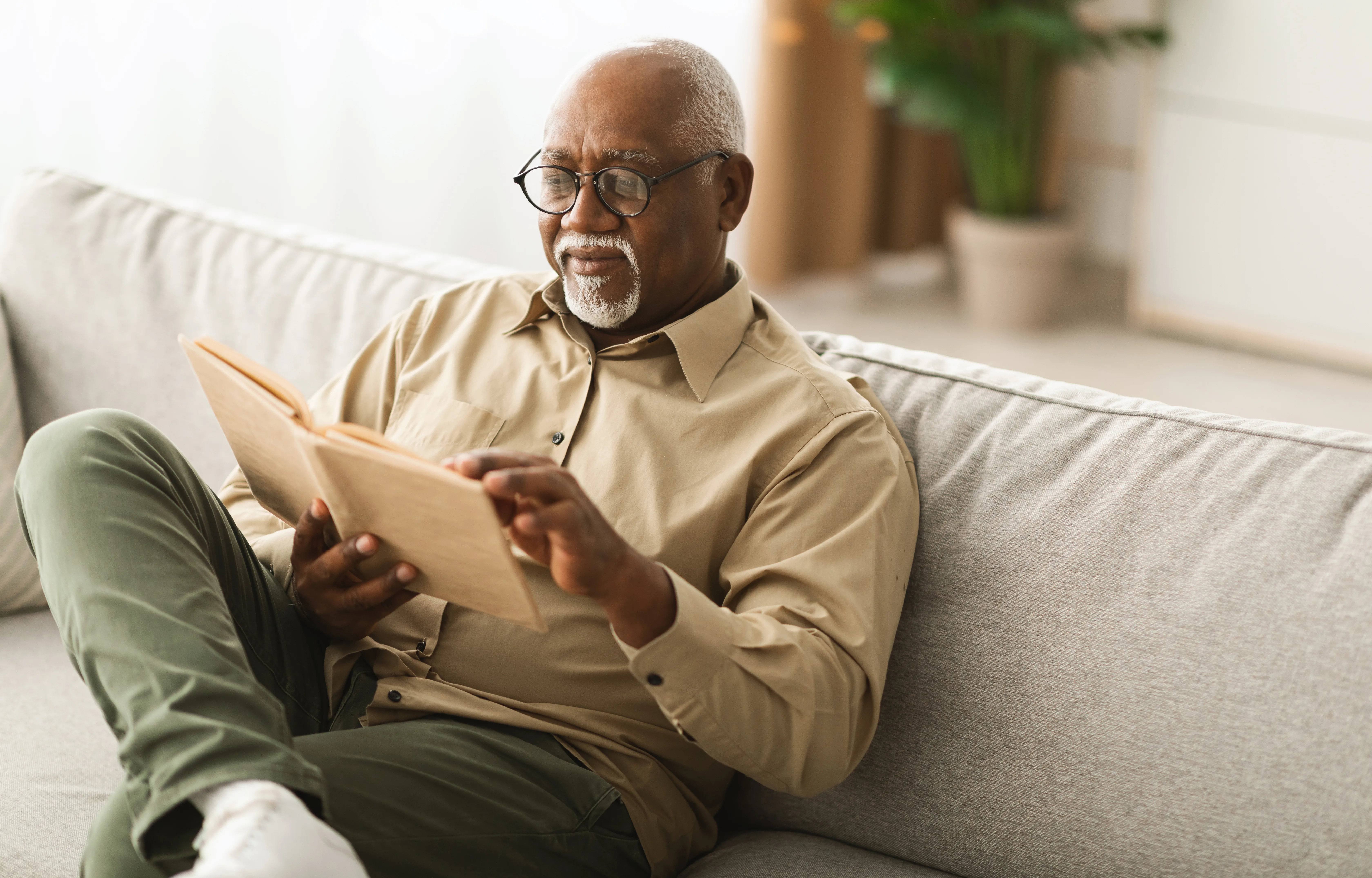 Mature Black Man Reading A Paper Book Relaxing Sitting On Sofa At Home, Wearing Glasses. Senior Gentleman Enjoying Reading New Novel On Weekend. Retirement Leisure And Lifestyle Concept