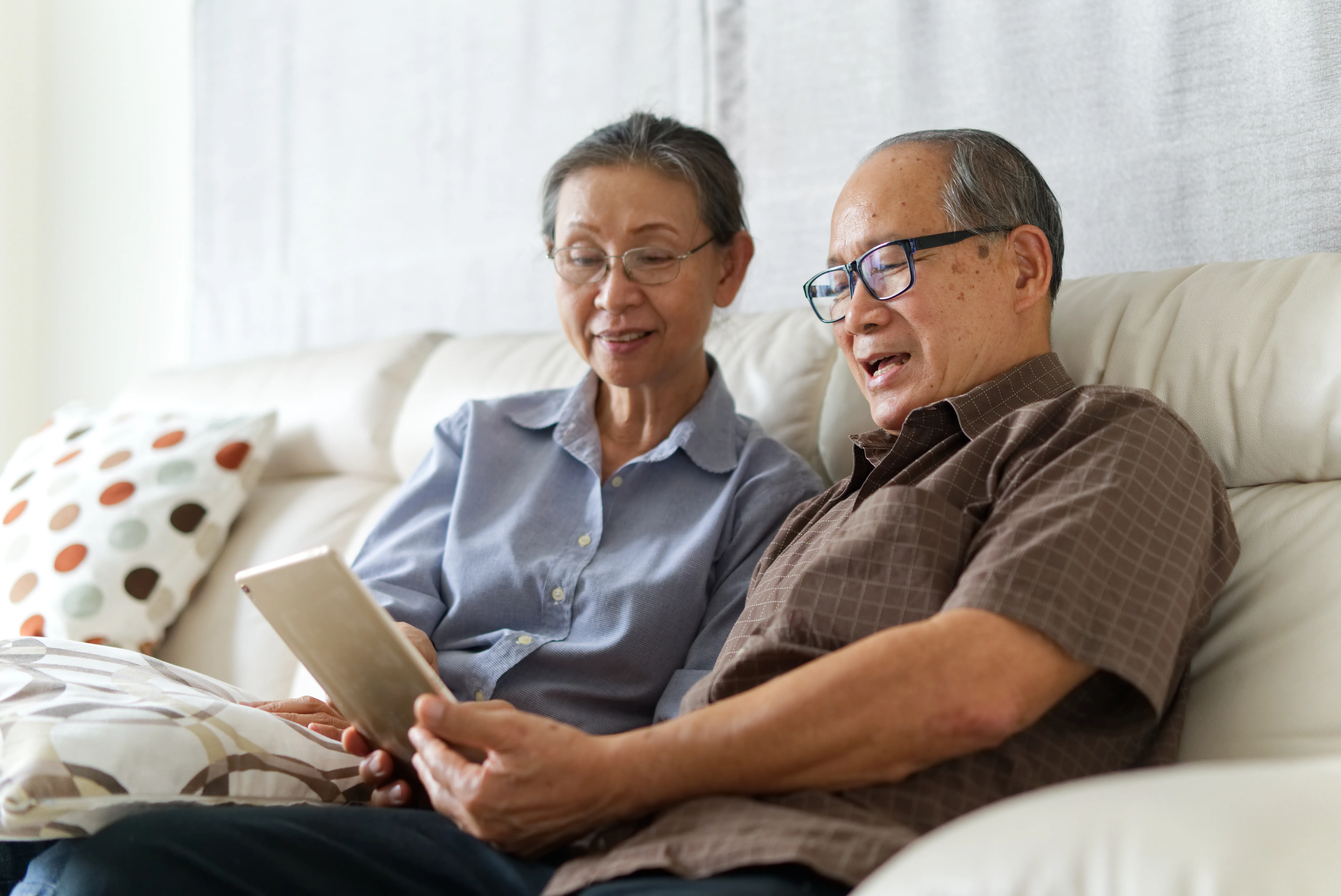 Senior couple sitting on sofa in home playing tablet and relaxing together. They are smiling and enjoy to spend their time together with happiness. Happy retirement life concept.
