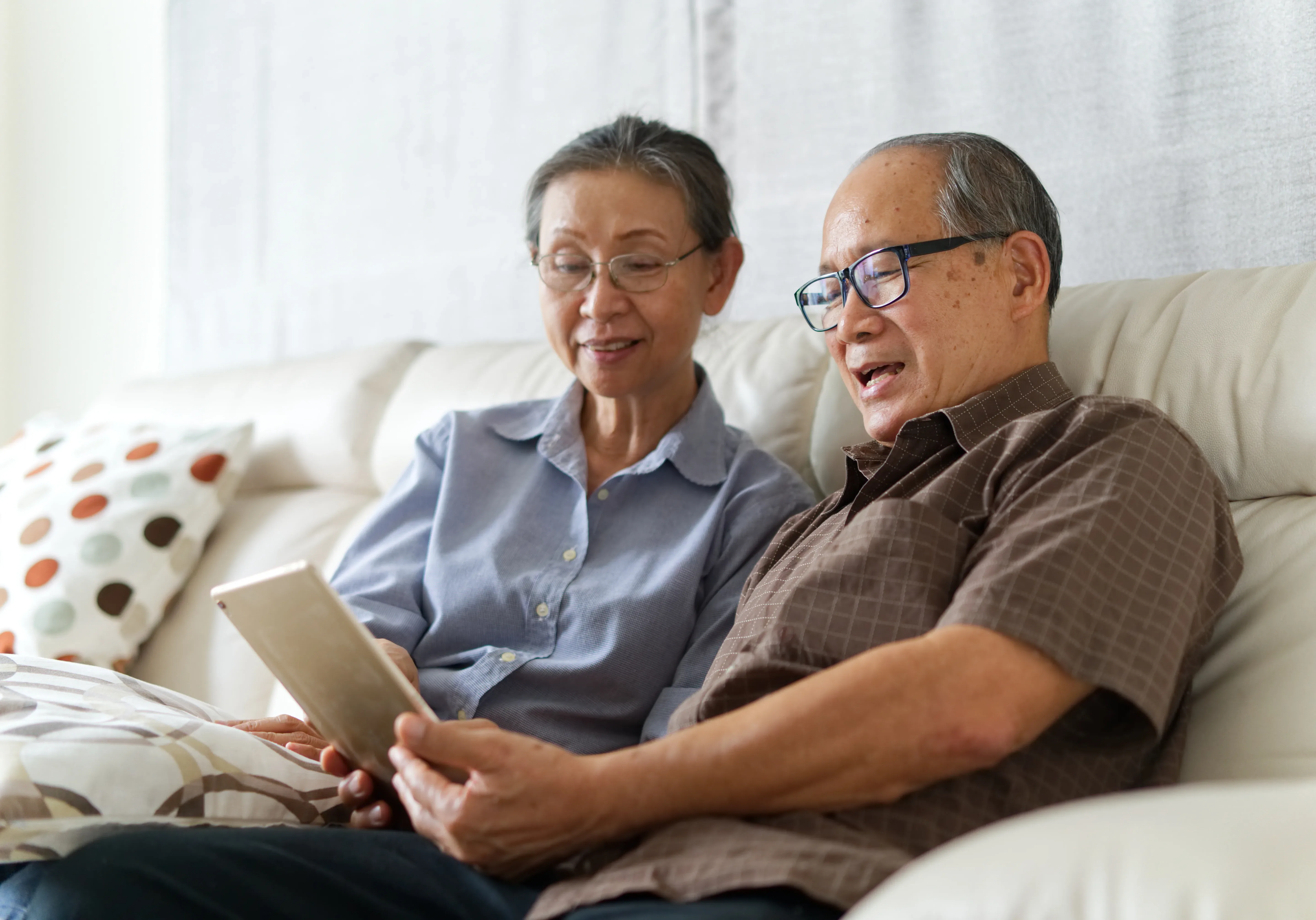 Senior couple sitting on sofa in home playing tablet and relaxing together. They are smiling and enjoy to spend their time together with happiness. Happy retirement life concept.